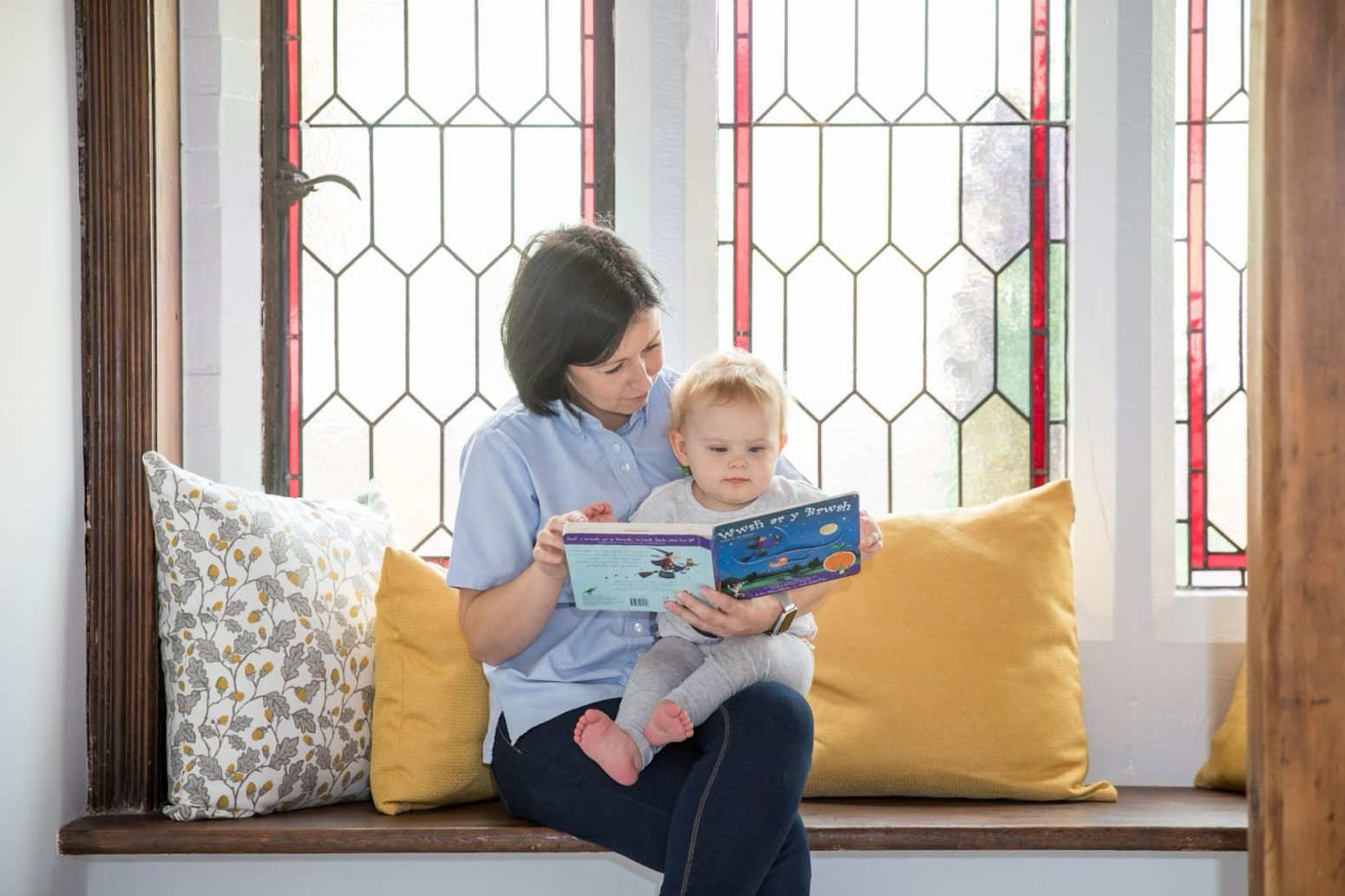 Acorns Nurseries An adult, with a caring smile, reads a book about space to a child on a bench with yellow cushions, in front of stained glass windows, creating a moment of cherished childcare amidst the vibrant colors.